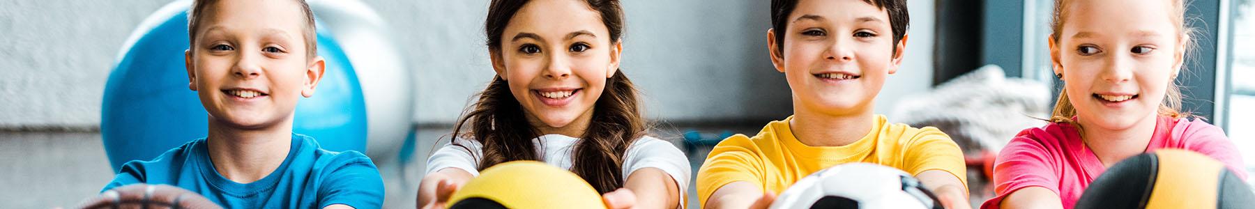 photo of 4 kids sitting on a bench smiling and holding out a different sports ball